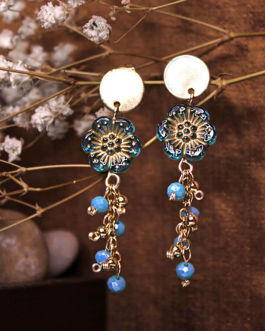 Gold plated stainless steel circle stud earrings with transparent light blue flower beads with gold outline detail. Dangling earrings with chain and blue faceted bead accent.